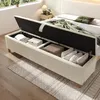 A beige upholstered storage bench with its lid open, revealing stored items like folded blankets and pillows, placed in a bedroom setting next to a bed and a nightstand with a lamp. Anteprima dell’immagine prodotto