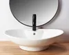 A white vessel sink with a black faucet is shown in a bathroom setting, with a round mirror in the background and a wooden surface in the foreground. Náhled obrázku produktu