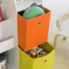 A close-up view of a white bookshelf with two fabric storage bins, one orange and one yellow, filled with toys and a plush toy. Produktbild-Vorschau