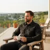 A man sits on a patio chair holding a mug of coffee, looking out at a scenic view. Produktbild-Vorschau