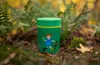 A green thermos with a cartoon character on it, placed on the forest floor surrounded by foliage. Produktbild-Vorschau