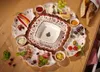 A collection of Villeroy & Boch Toy's Delight serving plates arranged on a wooden table, filled with various festive foods like cheese, grapes, cured meats, chili peppers, and bread. Anteprima dell’immagine prodotto