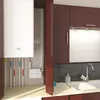 A white boiler unit is mounted on a wall above pipes and a small white box. To the right, a kitchen counter with a sink and faucet is visible, with dark tiled backsplash and dark cabinets above. Anteprima dell’immagine prodotto