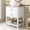 A white bathroom vanity with a ceramic sink and wooden countertop, featuring two drawers and an open shelf below, stocked with various bottles and jars. The background shows a tiled wall and a wall-mounted light fixture. Produktbild-Vorschau