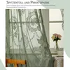 A close-up view of green lace curtains with floral patterns, partially covering a window. A vase with flowers and a teacup are visible on a table. Produktbild-Vorschau