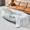 A plush, light gray pet bed with a white pillow and a matching blanket is placed on a light gray floor in front of a brown leather sofa and a wooden dresser. Produktbild-Vorschau