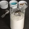 A close-up of white powder in a glass container with a metal scoop inside, next to three small white ramekins containing blue and colorful sprinkles, and another metal scoop on the side. Produktbild-Vorschau