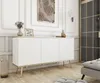 A white sideboard with gold legs is placed in a room with white paneled walls and a wooden floor. Decorative items are placed on top of the sideboard. Produktbild-Vorschau