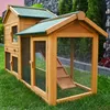 A wooden rabbit hutch with a wire mesh enclosure is set up on a green lawn, with a stone wall and trees in the background. Produktbild-Vorschau