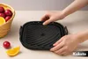 Hands holding a black silicone air fryer liner with a basket of fruit and an orange slice in the background. Náhled obrázku produktu