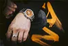 A person's arm wearing a watch and ring, with a black background featuring orange graffiti. Produktbild-Vorschau