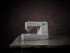 A gray Singer sewing machine is placed on a textured table against a dark, mottled background. Náhled obrázku produktu