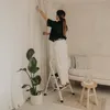 A woman is standing on a stepladder and adjusting a white curtain in a room with a sofa and a plant. Podgląd zdjęcia produktu