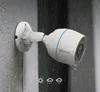 A white EZVIZ outdoor security camera is mounted on a textured gray wall, with water droplets on the camera and wall, suggesting it is raining. Voir les images du produit