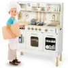 A child dressed as a chef stands next to a wooden toy kitchen. Produktbild-Vorschau