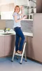 A woman is standing on a step stool in a modern kitchen, reaching into a cabinet. Náhled obrázku produktu
