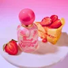 A bottle of perfume and a slice of strawberry cheesecake are displayed on a white marble stand against a pink gradient background. Podgląd zdjęcia produktu