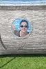 A young girl wearing swimming goggles smiles through a circular window in the side of a pool. The pool has a stone-like texture. The bottom of the image shows green grass. Produktbild-Vorschau