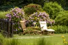 Two wooden deckchairs are set up on a green lawn in front of a large flowering bush with purple and pink blossoms. A small table between the chairs holds drinks. Produktbild-Vorschau