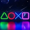 A neon sign displaying the PlayStation button symbols (triangle, circle, cross, square) against a starry night sky background with planets and shooting stars. The sign is reflected on a wet surface below. Produktbild-Vorschau