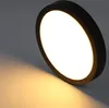 A close-up shot of a round, black LED ceiling light with a white diffuser, casting a warm yellow light onto a gray surface. Produktbild-Vorschau