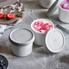 A set of white ice cream containers with gray lids, some filled with colorful ice cream, arranged on a gray surface with strawberries and blueberries nearby. Produktbild-Vorschau
