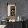 A mirror with a wooden frame is mounted on a brick wall above a dark wooden dresser. The dresser is decorated with various items, including plants, a lamp, and decorative objects. A table is partially visible in the foreground. Náhled obrázku produktu