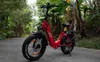 A red electric fat tire bicycle is parked on a paved path surrounded by lush green foliage and trees. Produktbild-Vorschau