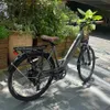 An electric bicycle is parked outdoors next to a planter with lush greenery. Produktbild-Vorschau