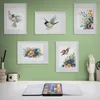A watercolor painting set is displayed on a desk with several framed watercolor paintings of birds and flowers on a green wall behind it. Podgląd zdjęcia produktu