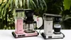 Two Moccacoffee makers are displayed on a white surface with a blurred green leafy background. Produktbild-Vorschau