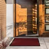A red doormat is placed on a concrete walkway in front of an open wooden door. Produktbild-Vorschau