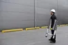 A man in a hard hat carries a folded aluminum telescopic ladder in front of a large gray building. Náhľad obrázku produktu