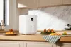 A white Xiaomi Mi Smart Air Fryer is placed on a wooden kitchen counter, with food items arranged around it. Podgląd zdjęcia produktu