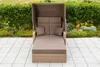 A brown wicker outdoor lounge chair with a canopy, set on a tiled patio with a green hedge and flowers in the background. Produktbild-Vorschau