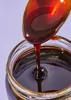 A spoon is lifting a viscous, dark brown liquid from a glass jar. Produktbild-Vorschau