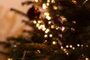A close-up of a Christmas tree ornament shaped like a candy cane, hanging among the branches and lights of a decorated Christmas tree. Produktbild-Vorschau