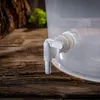 Close-up of a white plastic spigot attached to a transparent plastic container, set against a rustic wooden surface with blurred natural elements in the background. Anteprima dell’immagine prodotto