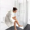 A woman in a white bathrobe is applying body scrub to her leg while sitting on a white fold-down shower seat in a modern bathroom. Produktbild-Vorschau