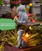 A split image showing a solar-powered cat statue in a garden setting. The top image shows the statue during the day with text about auto-charging, and the bottom image shows it lit up at night with text about auto-lighting. Produktbild-Vorschau