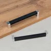 Two modern black and silver cabinet handles are displayed on a light wood surface and a light gray surface. Podgląd zdjęcia produktu