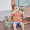 A young boy sits on steps, drinking from a green water bottle. A backpack and lunchbox are beside him. Produktbild-Vorschau