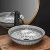 A decorative vessel sink with a black faucet and a gold-rimmed mirror in the background, placed on a wooden surface. Produktbild-Vorschau