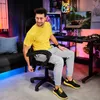 A man is sitting in a gaming chair in a room with gaming equipment. Produktbild-Vorschau