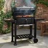A black charcoal grill with food cooking on it, placed on a stone patio next to a brick wall with ivy. Produktbild-Vorschau