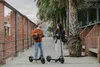 A man and a woman are standing with electric scooters on a paved path lined with trees and brick walls. Produktbild-Vorschau