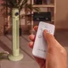 A hand holding a remote control in front of a tall, green tower fan. Podgląd zdjęcia produktu