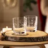 Two clear glasses with festive designs sit on a wooden slice next to a lit candle and a walnut. Produktbild-Vorschau