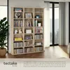 A light wood-colored bookshelf filled with books, CDs, DVDs, and decorative items, set against a backdrop of large windows with sheer curtains. Náhled obrázku produktu
