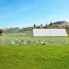 A large metal chicken coop with a white sunshade roof is set up in a grassy field with a few houses and trees in the background under a clear blue sky. Several chickens are visible inside the coop. Produktbild-Vorschau